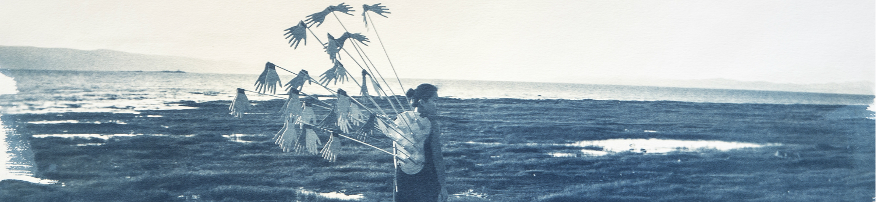 A cyanotype photo of a woman on a shoreline with an abstract art piece strapped to her back.