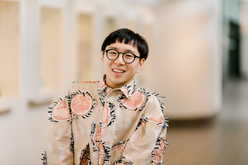 Sean Lee, a male-presenting East-Asian Chinese person smiles at the camera. He wears steampunk glasses that are green with gold frames, and has cropped hair with blunt bangs. He wears a beige shirt