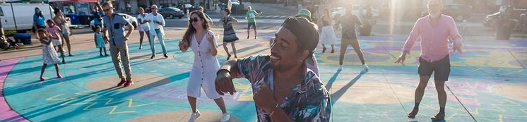 A group of people caught mid motion in a collective dance are spread across the frame. They stand on brightly coloured painted concrete outdoors