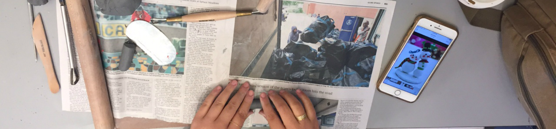 Two hands roll out clay on a newspaper. Clay sculpting tools are on the left of the table, while a cellphone is on the right.
