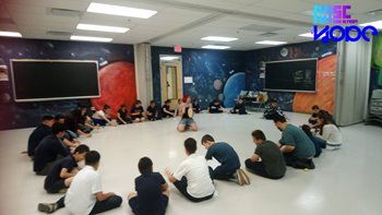 a woman with a tambourine sits in the middle of a large circle of men women and children in a room. The walls are painted with a mural of a dark sky with stars and planets.