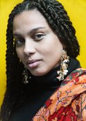 Paulina Anthony looks intensely at camera against a yellow background. Her hair is braided and she wears a black turtleneck with a colourful scarf on her left shoulder, and dangling earrings 