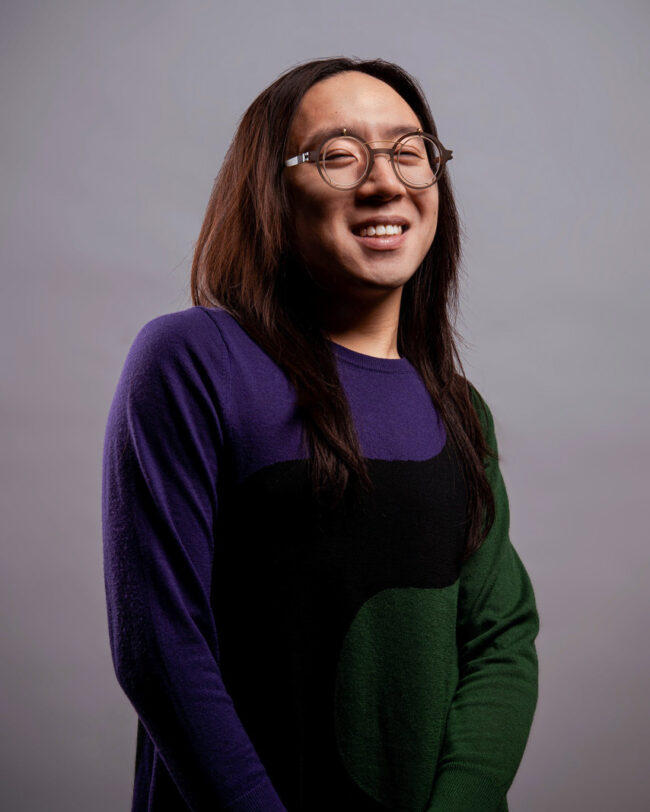 Sean Lee, a male-presenting East-Asian Chinese person smiles at the camera. He wears steampunk circle glasses that are brown with gold frames, and has wavy dark brown hair that pools at his shoulders. He wears a long sleeve dress that is green and purple with a thick winding black stripe that runs down the middle.