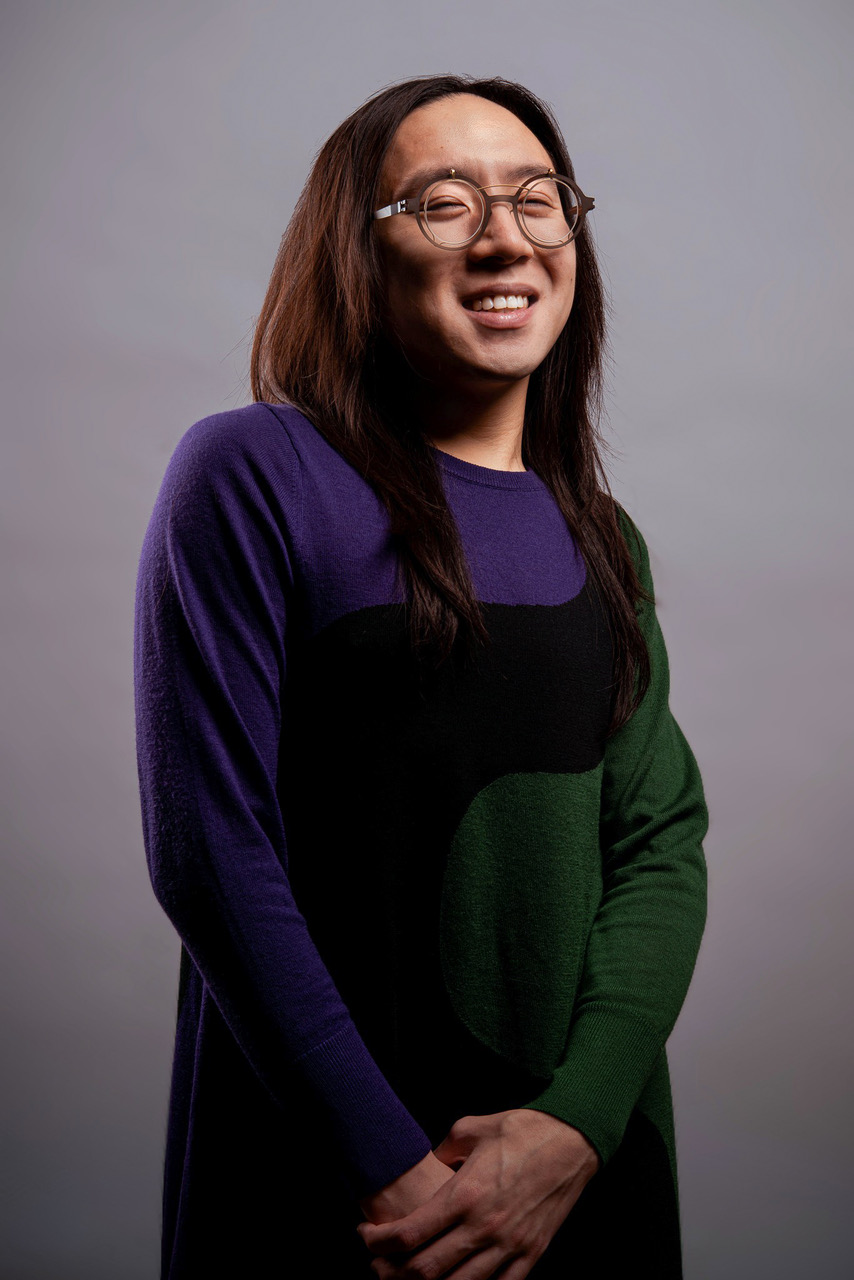 Sean Lee, a male-presenting East-Asian Chinese person smiles at the camera. He wears steampunk circle glasses that are brown with gold frames, and has wavy dark brown hair that pools at his shoulders. He wears a long sleeve dress that is green and purple with a thick winding black stripe that runs down the middle.