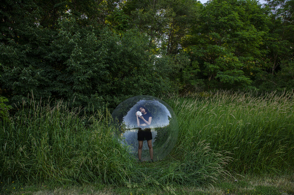 A person stands within a transparent round large balloon, on a field of grass