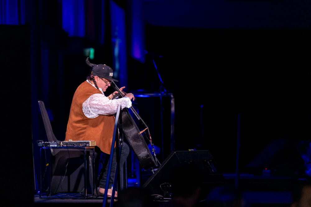 A photo of Cris Derksen playing cello on a dark stage