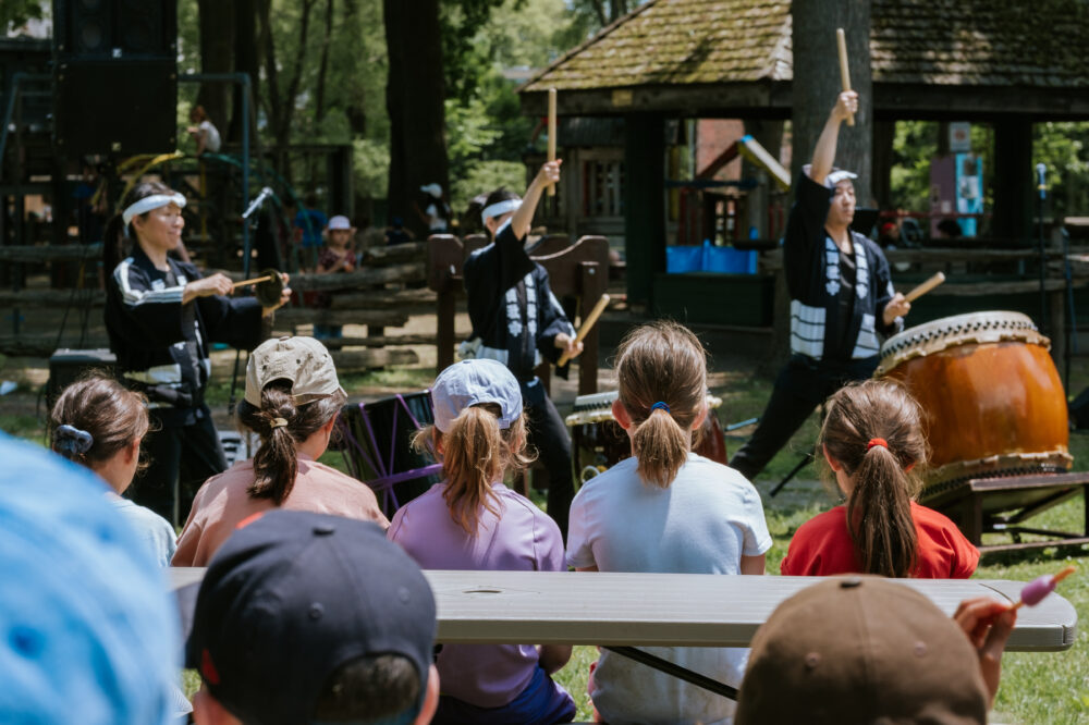 A group of children are seen from behind sitting at a picnic table at a park, watching a drumming performance.