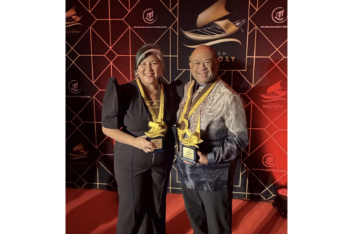 Ana Serrano and Michael Herrara pose in front of a step-and-repeat holding award trophies.