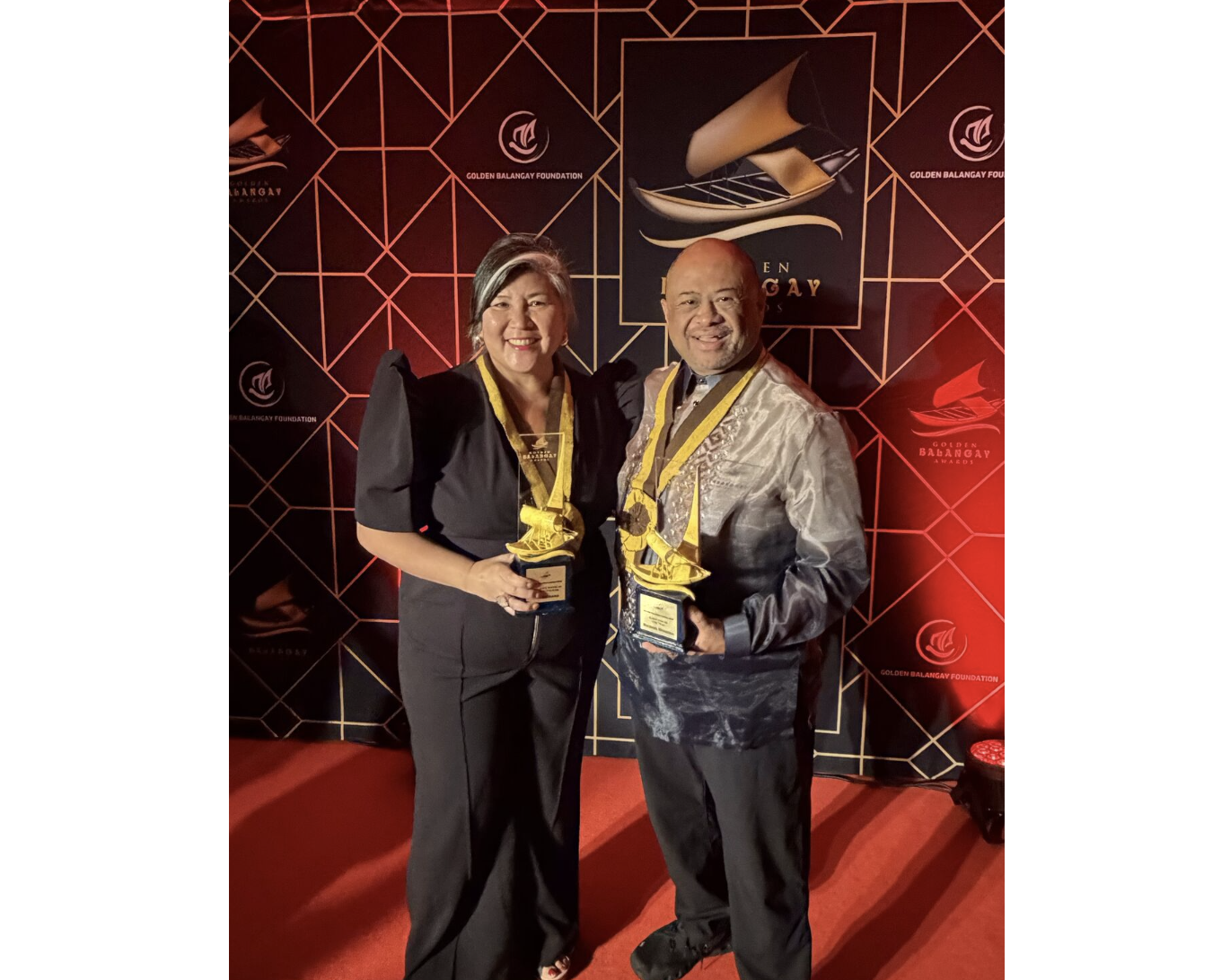 Ana Serrano and Michael Herrara pose in front of a step-and-repeat holding award trophies.